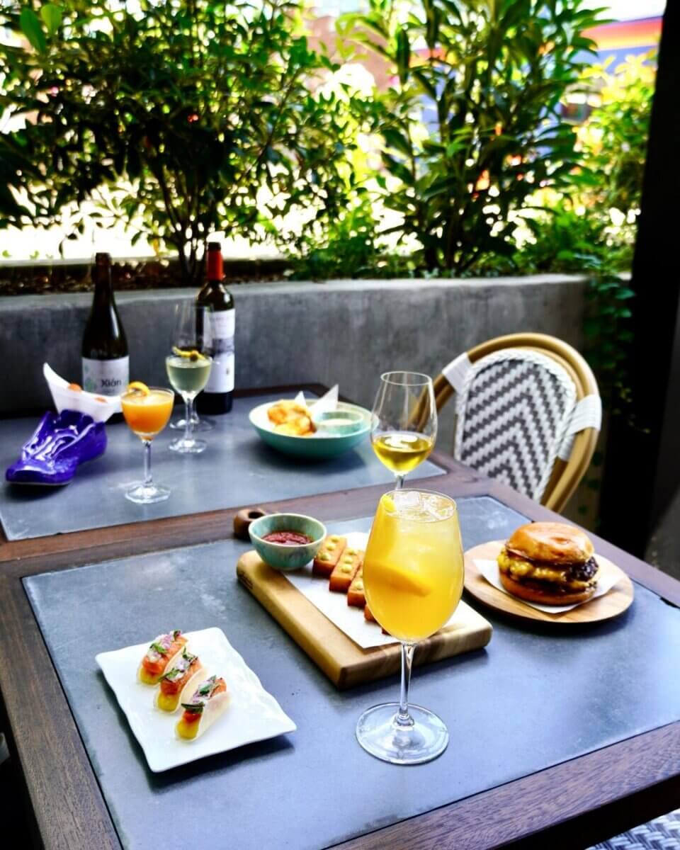 Nashville Steals, Deals & Announcements: July 2026 - 11 A restaurant patio table with wine, cocktails, and plates of sushi, bread, fries, and a burger—set for two with greenery in the background.
