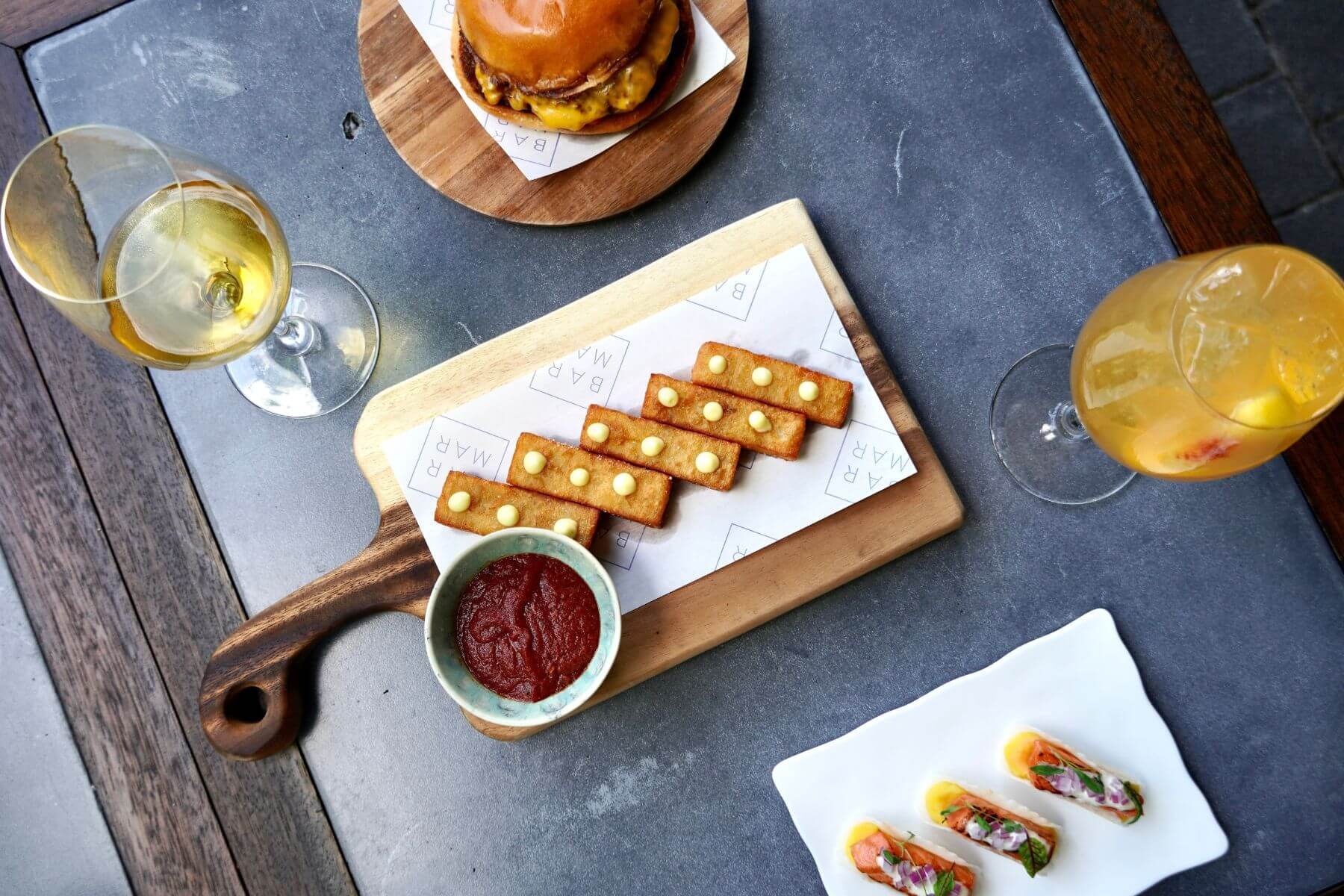 A tabletop with two glasses of white wine, a cheeseburger, a wooden board with rectangular crackers and dip, plus a small plate of appetizers with fish and garnish.