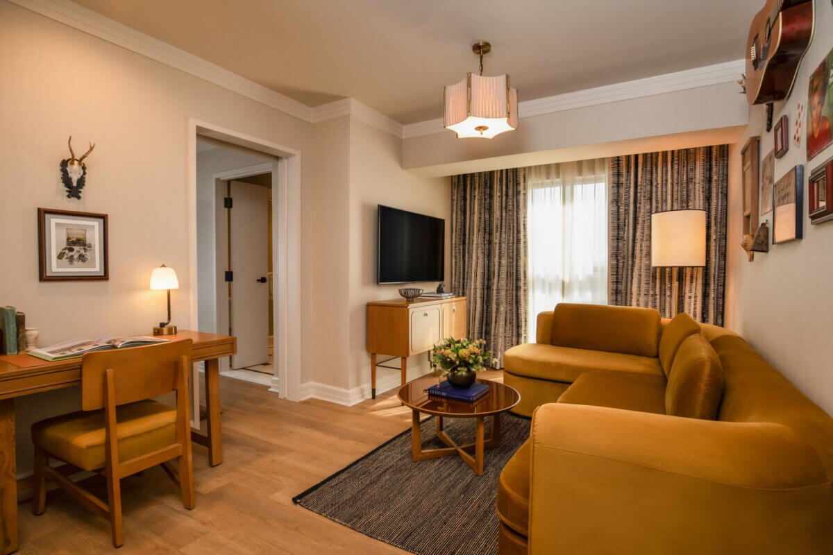 Nashville Steals, Deals & Announcements: July 2026 - 2 A living room with mustard yellow sofa, coffee table with flowers, TV on the wall, desk with chair and lamp, wood flooring, and art on the walls. Light enters through curtained window—explore Nashville Steals for similar July 2026 styles.