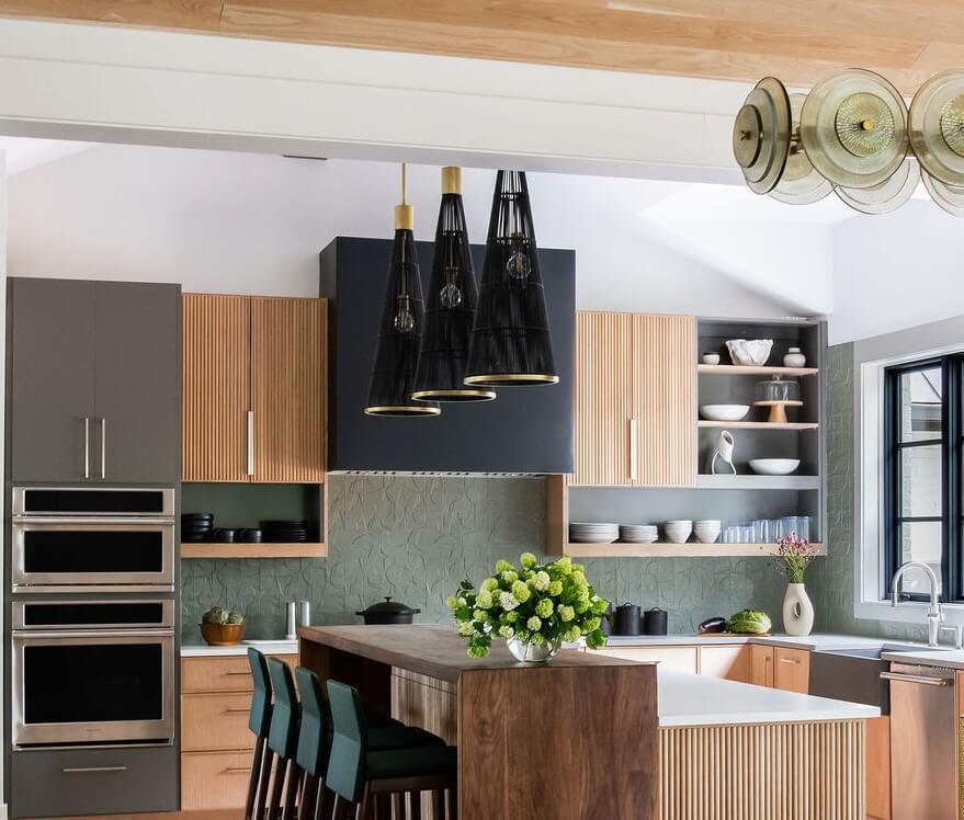 Modern kitchen with wood and dark cabinets, an island with green chairs, pendant lights, built-in ovens, open shelves with dishes, and a vase of green flowers—perfect inspiration if you’re looking to hire an interior designer.