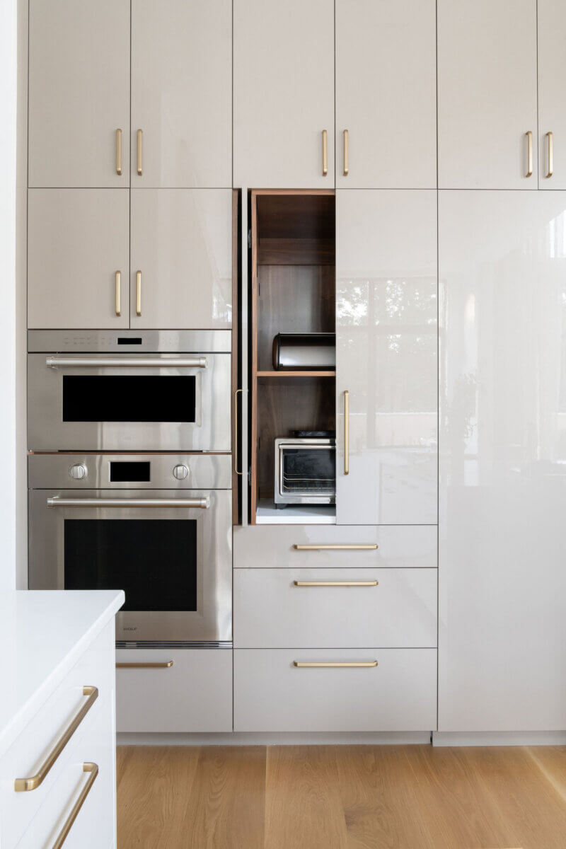 Modern kitchen with glossy white cabinets, gold handles, stainless steel built-in oven, microwave, and an open section revealing a toaster and bread box—perfect inspiration if you plan to hire an interior designer for your home.