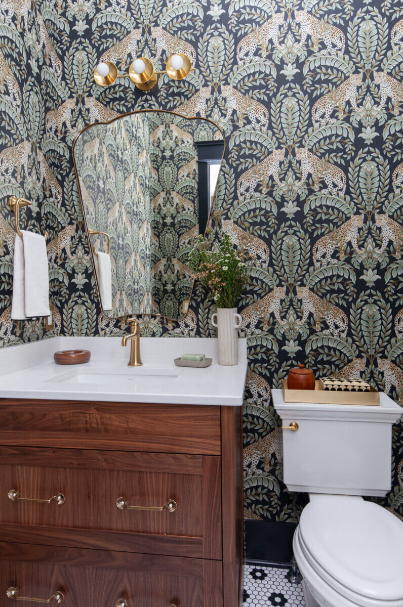A small bathroom designed by an interior designer features a wooden vanity, gold fixtures, a geometric mirror, patterned wallpaper with leopards and foliage, and a white toilet beside a vase of flowers.