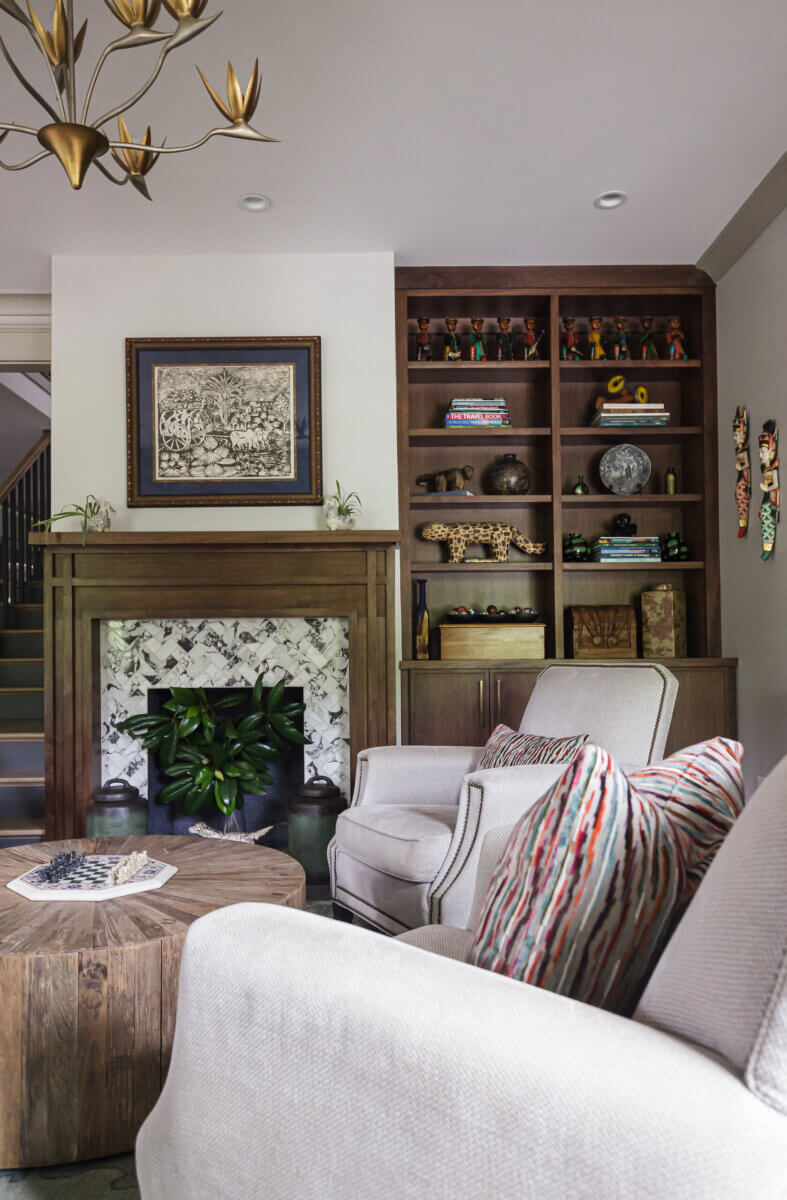 A living room with beige armchairs, a wooden coffee table, and a fireplace with a plant inside highlights how an interior designer can elevate your space. Built-in shelves and artwork above the mantel complete the refined interior design process.