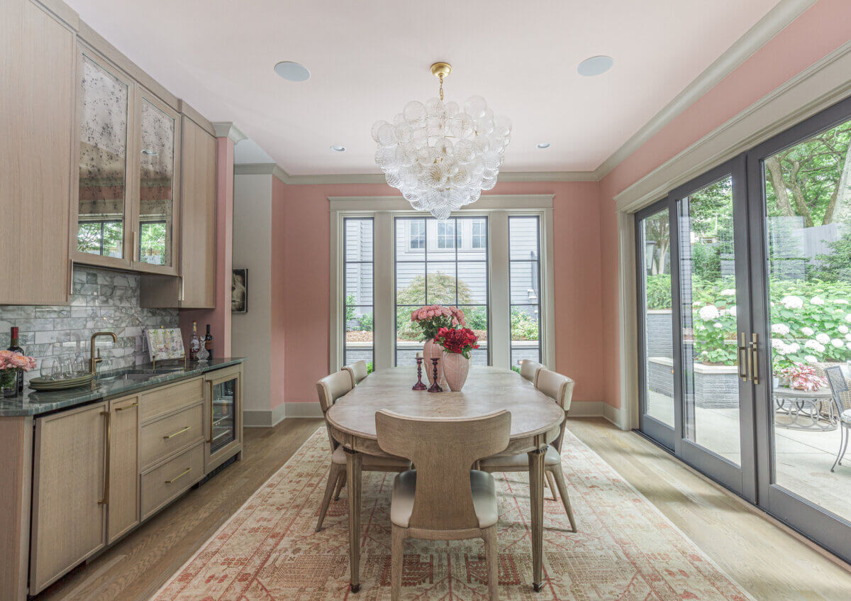 A dining room with a round wooden table, six chairs, a glass chandelier, built-in cabinets, pink walls, and large windows overlooking a garden patio—perfect inspiration if you want to hire an interior designer for your own space.