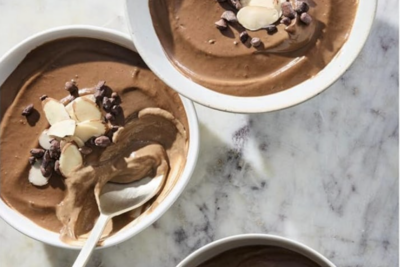 Three bowls of chocolate mousse topped with sliced almonds and chocolate chips are arranged on a marble surface. A spoon rests in one of the bowls.