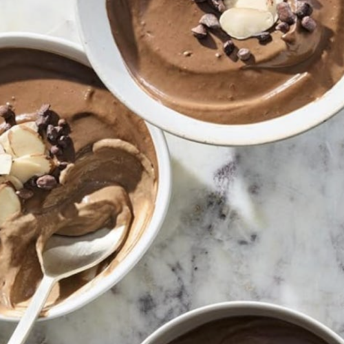RECIPE: Chocolate Protein Pudding - 3 Three bowls of chocolate mousse topped with sliced almonds and chocolate chips are arranged on a marble surface. A spoon rests in one of the bowls.