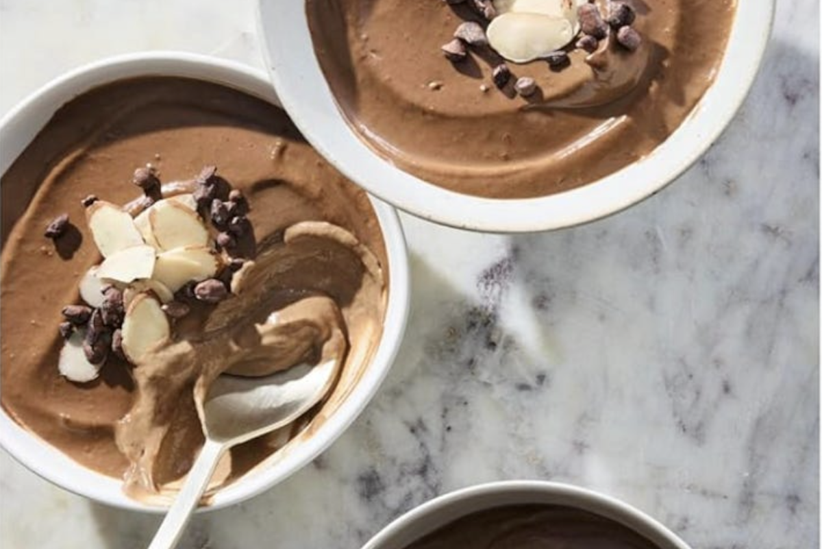 Three bowls of chocolate mousse topped with sliced almonds and chocolate chips are arranged on a marble surface. A spoon rests in one of the bowls.