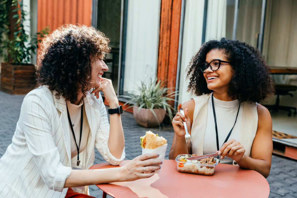 Not All Proteins Are Created Equal! Here's How to Eat for Your Goals - 3 Two women sit at an outdoor table, smiling and talking while eating lunch. One holds a wrapped sandwich; the other enjoys a balanced meal from a container with a fork.