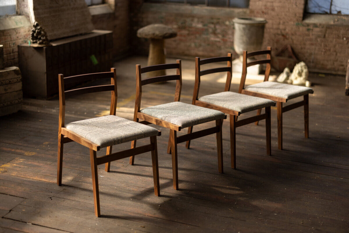 Turning Fallen Trees into Fine Furniture: The Art of Alabama Sawyer - 4 Four wooden chairs with light-colored cushioned seats are arranged in a line on a wooden floor in a sunlit, rustic room.
