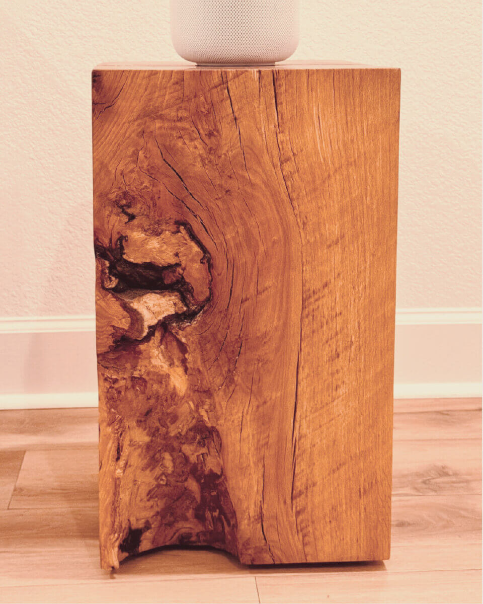 Turning Fallen Trees into Fine Furniture: The Art of Alabama Sawyer - 6 A rectangular wooden block with a natural knot and textured grain stands on a hardwood floor, partially supporting a white cylindrical object on top.