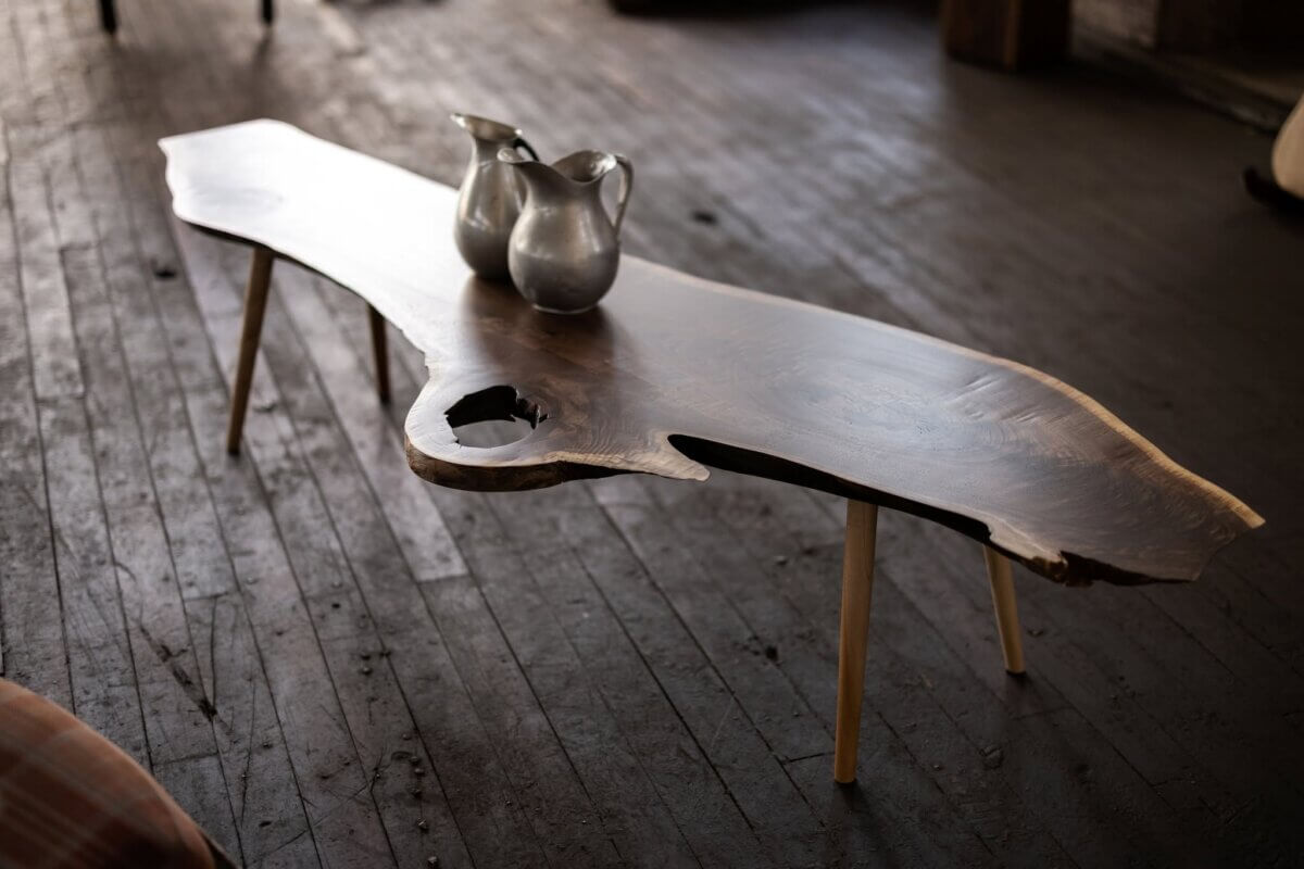 Turning Fallen Trees into Fine Furniture: The Art of Alabama Sawyer - 2 A rustic wooden coffee table with natural edges and tapered legs, holding two metallic pitchers, sits on a dark wooden floor.