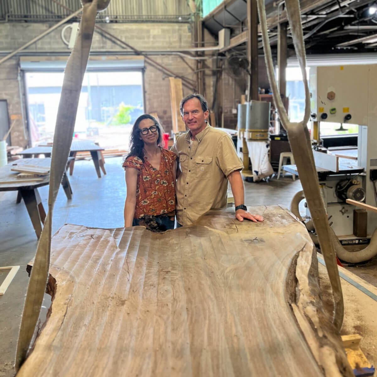 Turning Fallen Trees into Fine Furniture: The Art of Alabama Sawyer - 3 Leigh and Cliff Spencer stand behind a large wooden slab in a workshop, with various tools and equipment visible in the background at Alabama Sawyer.