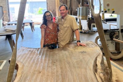 Leigh and Cliff Spencer stand behind a large unfinished wooden table in a woodworking shop with various tools and equipment in the background at Alabama Sawyer.