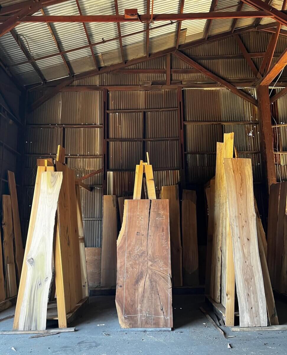 Turning Fallen Trees into Fine Furniture: The Art of Alabama Sawyer - 8 Tall, rough-cut wooden planks stand vertically inside a dimly lit warehouse with a metal roof and corrugated metal walls at Alabama Sawyer.