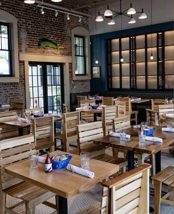 Turning Fallen Trees into Fine Furniture: The Art of Alabama Sawyer - 9 A modern restaurant dining area at Magnolia Point with wooden tables and chairs by Alabama Sawyer, place settings, brick walls, and pendant lighting. A fish decoration is mounted above double doors.