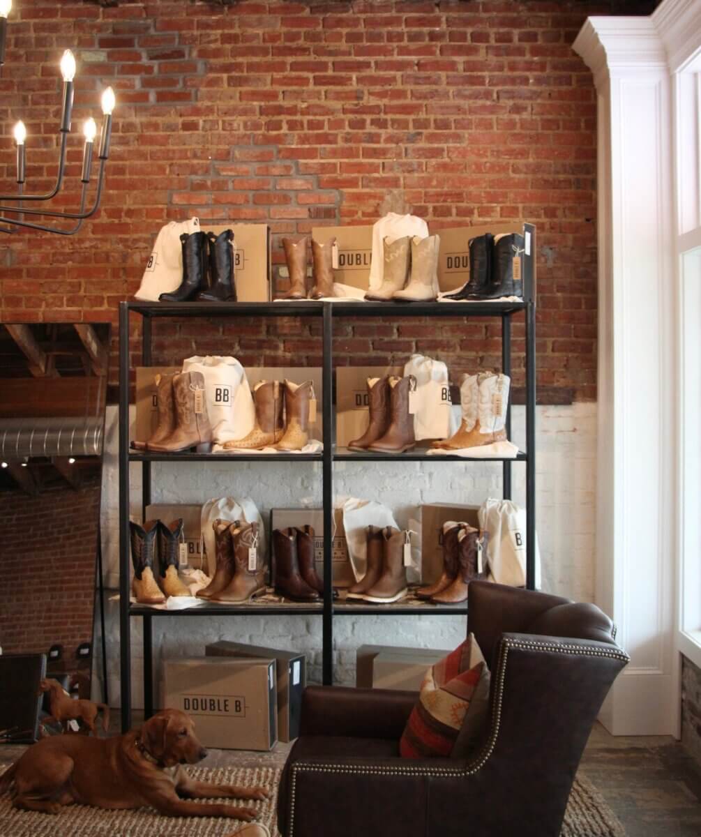 This Mississippi-Based Boot Company Should Be on Your Radar - 7 A shelving unit displays cowboy boots and Double B boxes against an exposed brick wall, reflecting classic Western style; a brown dog lies on the floor beside a leather chair.