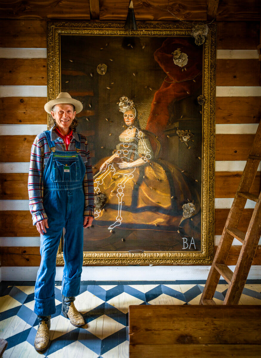 Inside Alabama Artist Butch Anthony's "Museum of Wonder" - 6 A man in overalls and a hat stands smiling beside a large, framed portrait with bullet holes on a wooden wall inside Butch Anthonyβs quirky Museum of Wonder, its patterned floor tiles adding to the roomβs rustic charm.