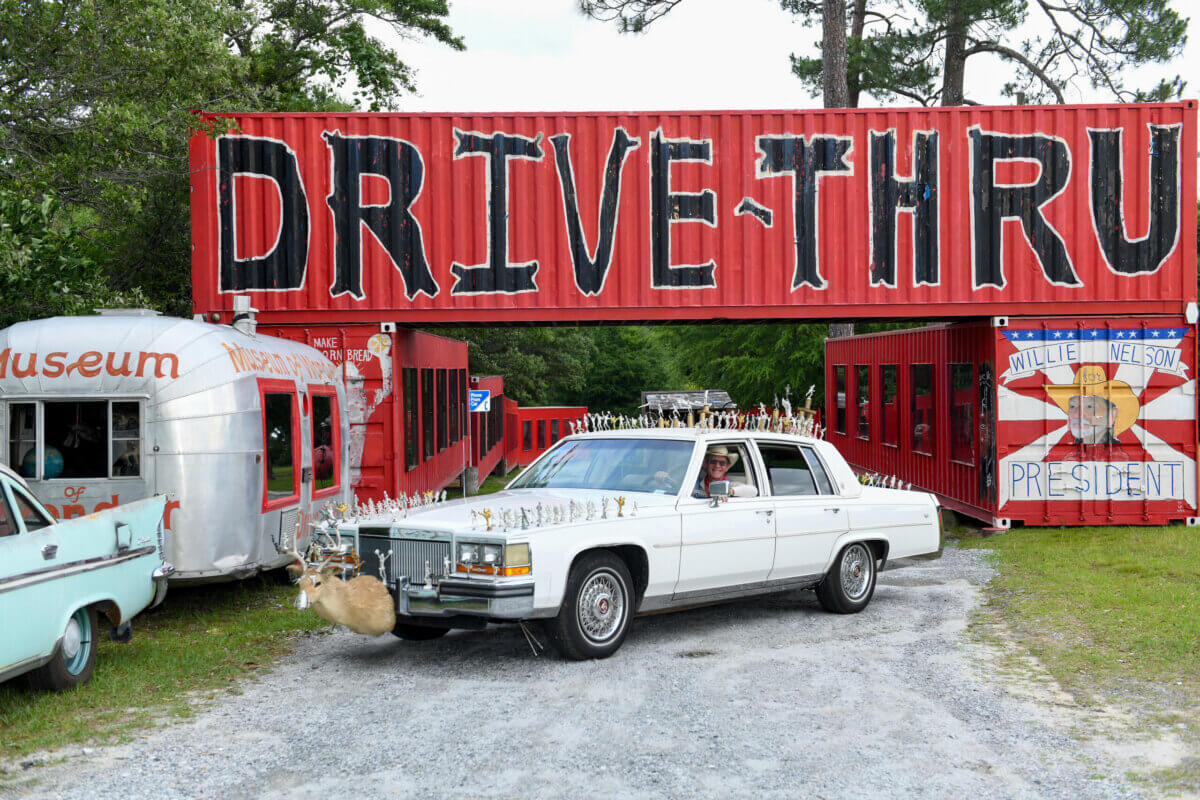 Inside Alabama Artist Butch Anthony's "Museum of Wonder" - 4 A white vintage car with antlers on the hood is parked in front of a red "Drive-Thru" sign made from shipping containers at Butch Anthony's Museum of Wonder, with a trailer and mural visible nearby.