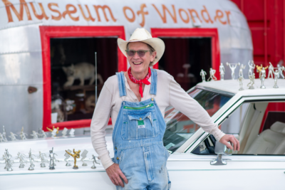 A man in overalls and a cowboy hat stands smiling next to a white car decorated with figurines, in front of Alabama's Museum of Wonder.