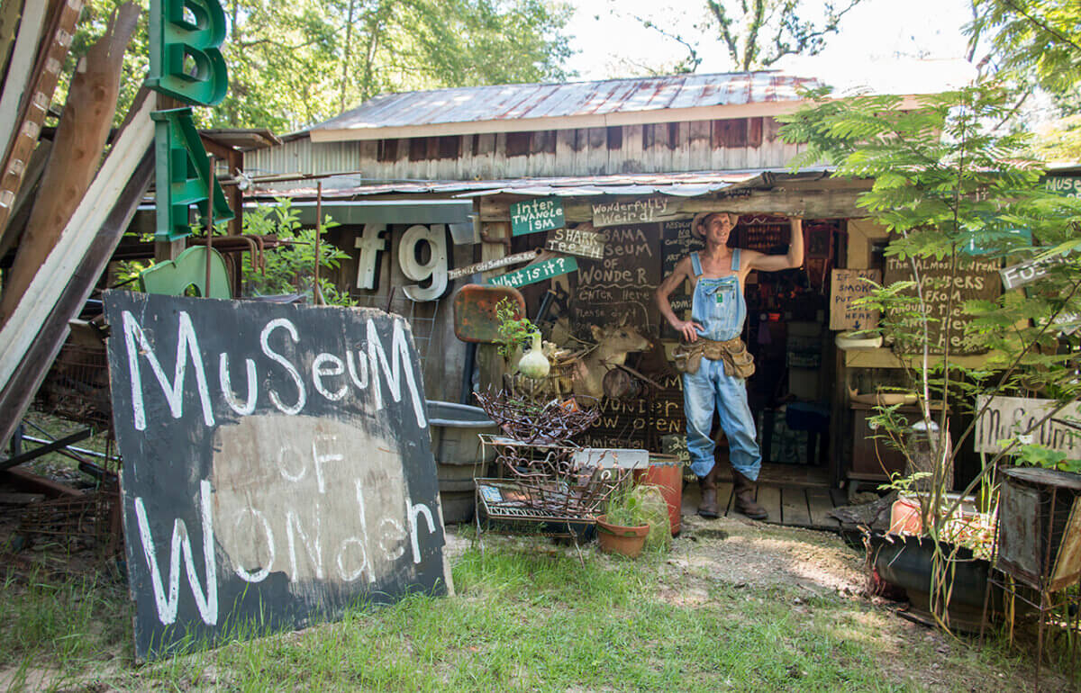 Inside Alabama Artist Butch Anthony's "Museum of Wonder" - 2 A person in overalls stands at the entrance of the Museum of Wonder, Butch Anthonyβs rustic haven, surrounded by handmade signs, plants, and eclectic objects.