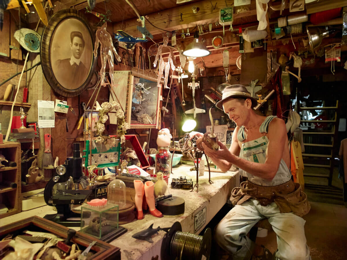 Inside Alabama Artist Butch Anthony's "Museum of Wonder" - 3 Butch Anthony, in a hat and overalls, works at a cluttered workbench in the dimly lit Museum of Wonder, surrounded by tools, trinkets, and a vintage portrait.