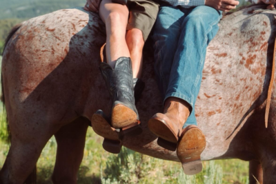 Two people sit side by side on a horse, showing only their legs and boots; one wears black cowboy boots, the other brown boots and jeans.