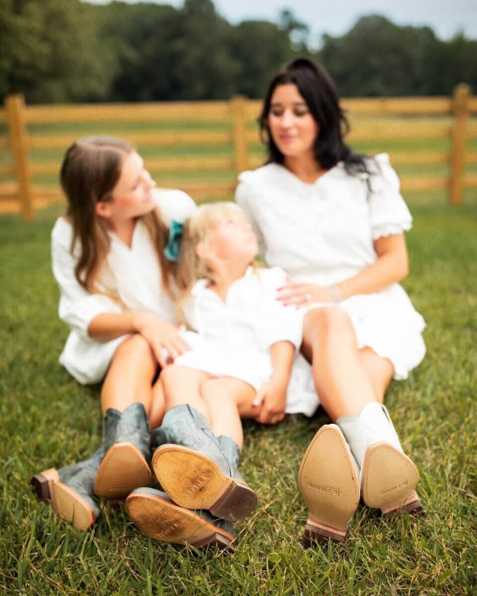 This Mississippi-Based Boot Company Should Be on Your Radar - 4 Three people in classic Western style outfits and boots sit on grass before a wooden fence, trees behind them. Their faces are slightly blurred and their boots face the camera.