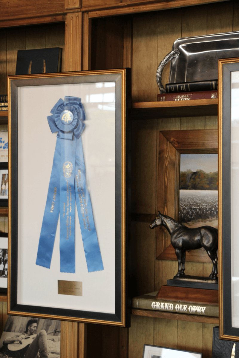 This Mississippi-Based Boot Company Should Be on Your Radar - 6 A framed blue ribbon displayed on a wooden shelf, next to a silver purse, a horse statue, and booksβincluding one labeled "Grand Ole Opry"βevokes classic Western style and deep Mississippi roots.