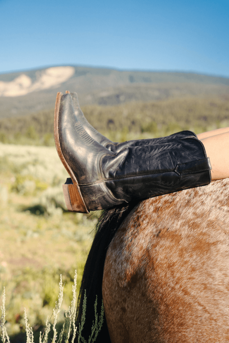This Mississippi-Based Boot Company Should Be on Your Radar - 5 Person wearing black cowboy boots from Double B Boot Company rests feet on the back of a brown horse in an open landscape with hills and blue sky, embodying true Western Style.