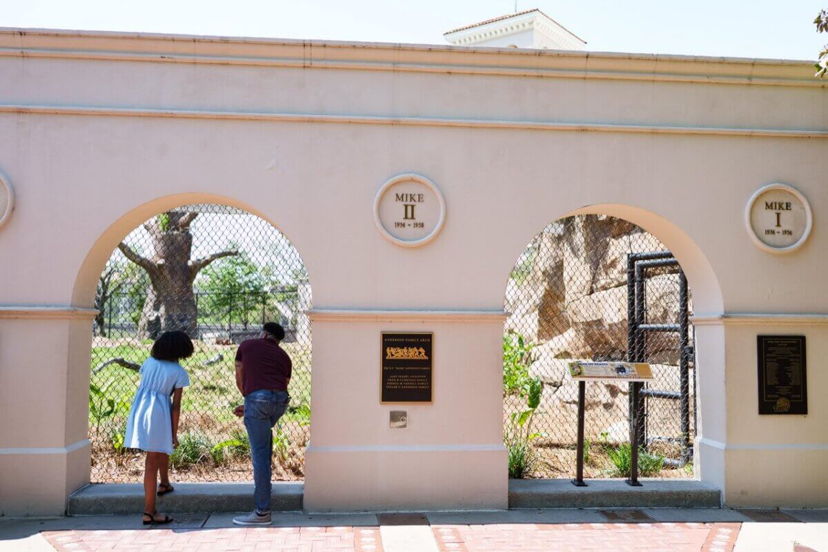 Two people stand and look through arched openings in a wall with commemorative plaques, observing an enclosed outdoor exhibit.
