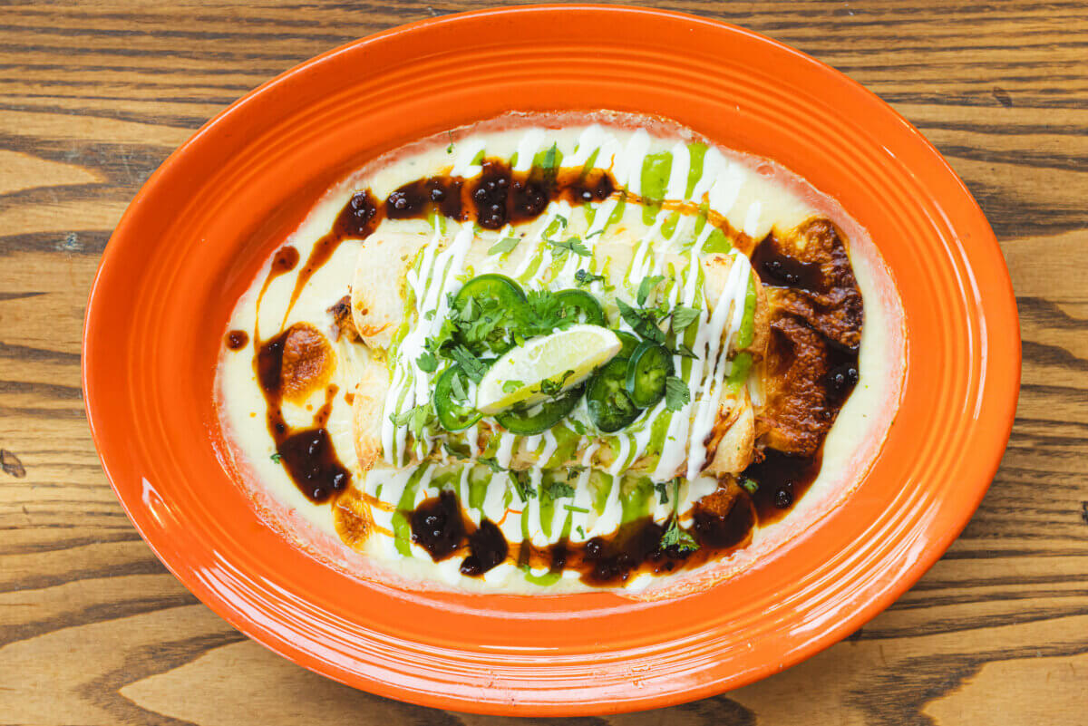 An orange plate with a baked cheese dip topped with sliced jalapeños, cilantro, lime wedge, and drizzles of green and dark sauces.