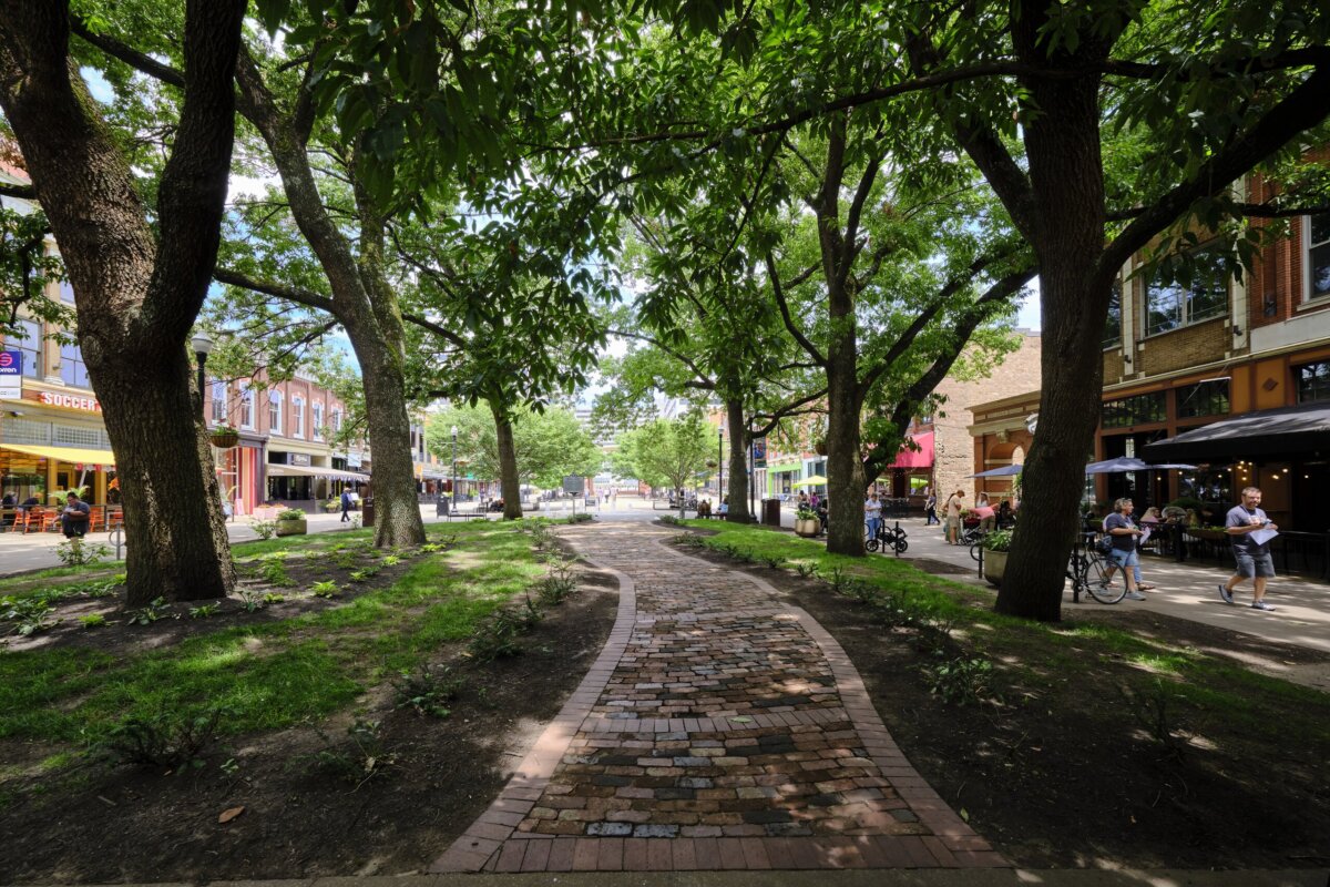 A brick path winds through a shaded park area with large trees, surrounded by shops, restaurants, and people enjoying a sunny day in one of the top SEC towns.