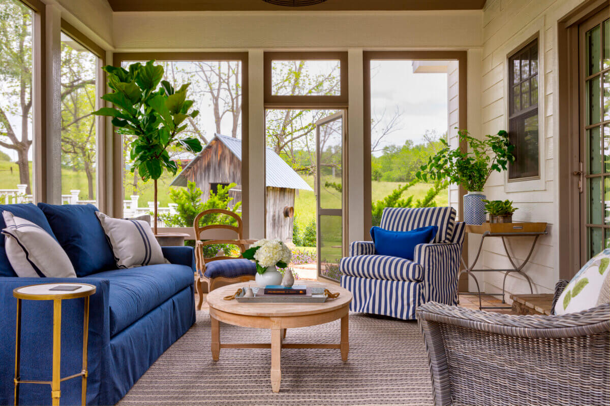 Daniel Keeley on Designing Outdoor Spaces That Actually Get Used - 13 A screened-in porch by Daniel Keeley features a blue sofa, striped armchair, wicker chair, round wooden coffee table, potted plants, and a view of the green yard with a shedβcreating a relaxed yet sophisticated style.
