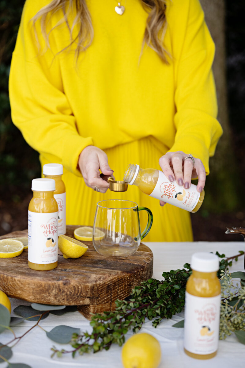 Trusting Her Gut: Meet the Charlotte Woman Making Healthy Habits Easier - 9 A mom entrepreneur in a yellow outfit pours juice from a bottle into a glass mug, surrounded by lemons, bottles, and lush green foliageβcapturing the essence of a wellness brand on a wooden and white surface.