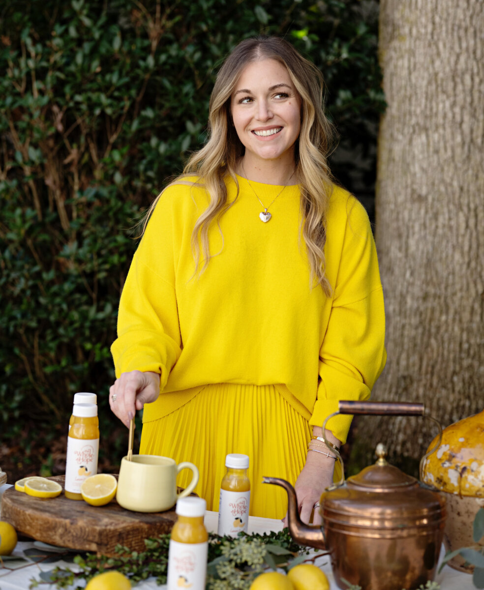 Trusting Her Gut: Meet the Charlotte Woman Making Healthy Habits Easier - 2 A mom entrepreneur in a bright yellow outfit stands at an outdoor table with lemons, bottled drinks, a teapot, and a yellow mug, smiling and stirring a drink for her wellness brand.