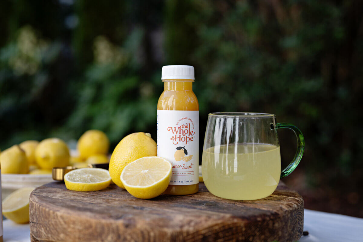 Trusting Her Gut: Meet the Charlotte Woman Making Healthy Habits Easier - 4 A bottle of The Whole Hope lemon juice, a glass mug of lemon juice, and fresh lemons are arranged on a wooden board outdoors.