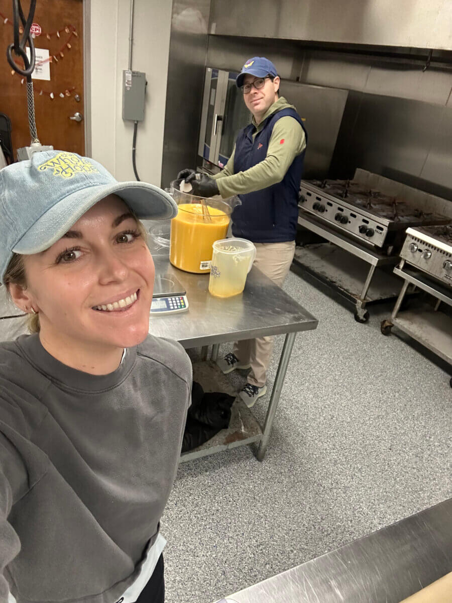Trusting Her Gut: Meet the Charlotte Woman Making Healthy Habits Easier - 5 Two people in a commercial kitchen prepare food on a metal table. One smiles at the camera, the other mixes something in a large yellow container. Cooking equipment is visible in the background.