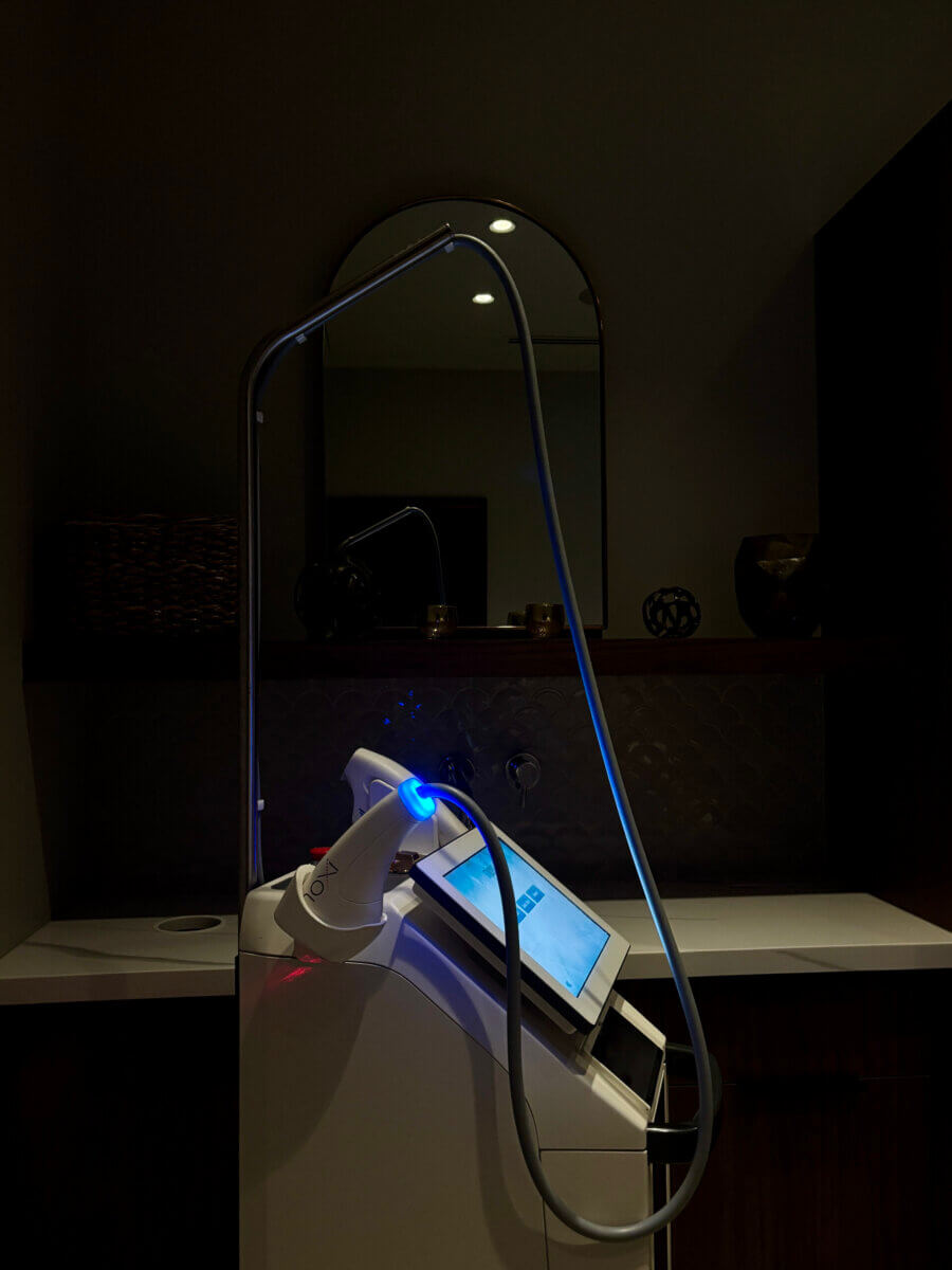 Yes, You Can Get Laser Treatments in Summer! Here's What to Know. - 3 A medical or cosmetic device with a touchscreen and flexible tube, illuminated by blue light, set in a dimly lit room with a counter and shelves in the background.