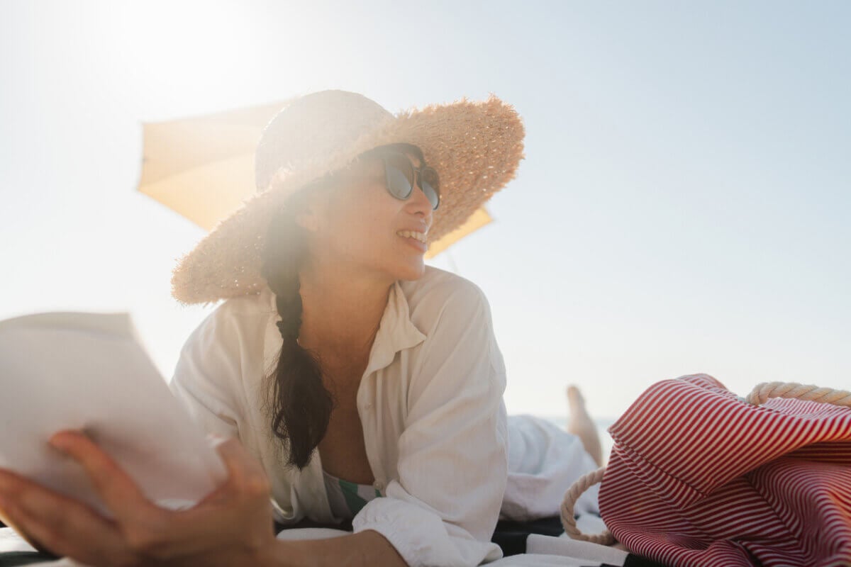 Woman in a sunhat and sunglasses lies on a beach towel, holding a book, with a striped bag and umbrella nearby on a sunny day—relaxing after researching the safest lasers for summer.