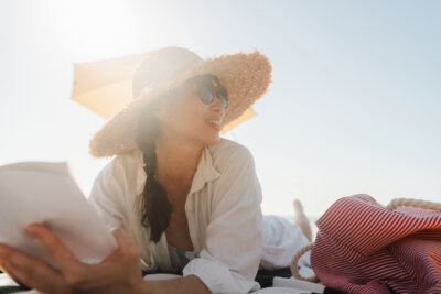 Woman in a sunhat and sunglasses lies on a beach towel, holding a book, with a striped bag and umbrella nearby on a sunny day—relaxing after researching the safest lasers for summer.