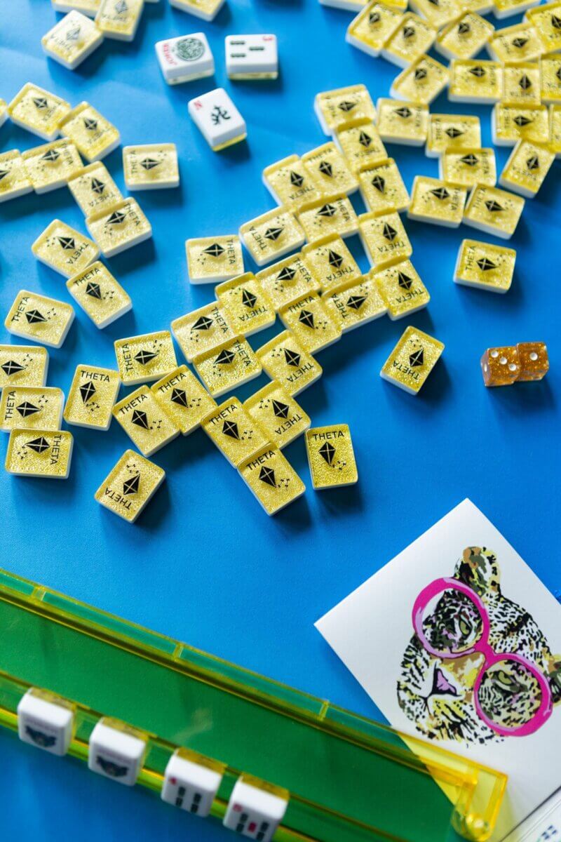 Yellow Mahjong tiles with "THETA" and dice are scattered on a blue surface, next to a green tile rack and a card featuring a leopard illustration—perfect decor for any Kappa Alpha Theta sorority house game night.