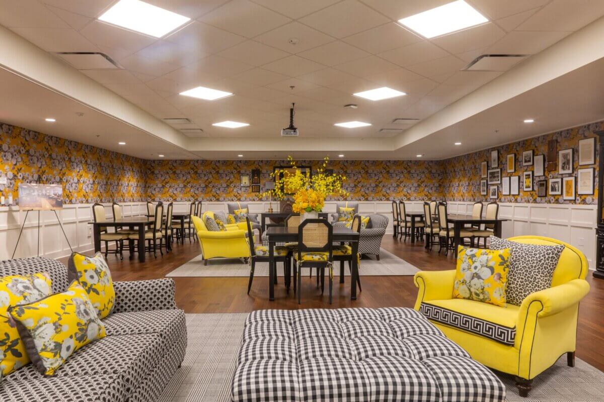 A spacious room in a Kappa Alpha Theta sorority house, featuring checkered and floral upholstered furniture, yellow accents, patterned wallpaper, framed pictures on the wall, and a central table with a yellow flower arrangement.