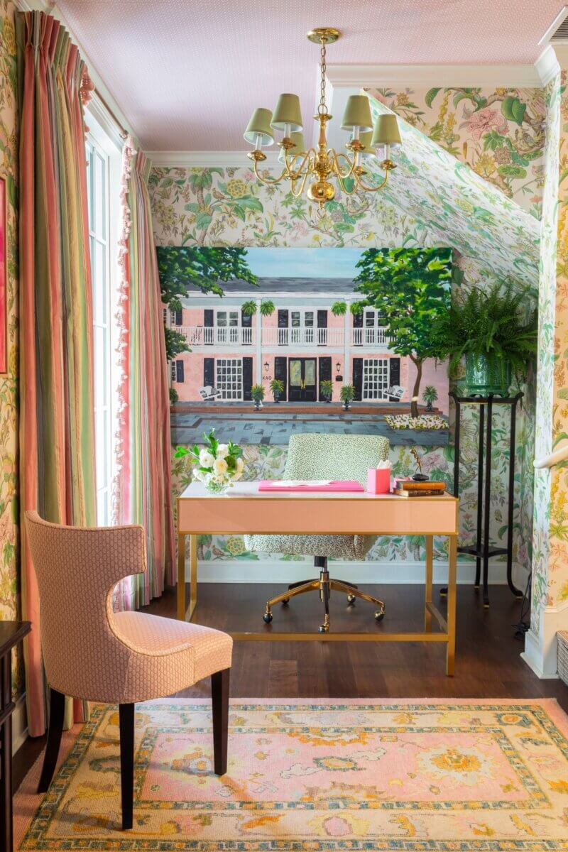 A home office with floral wallpaper, a wooden desk, pink chair, gold chandelier, and a mural of a large Kappa Alpha Theta sorority house on the back wall. A patterned rug covers the dark wood floor.