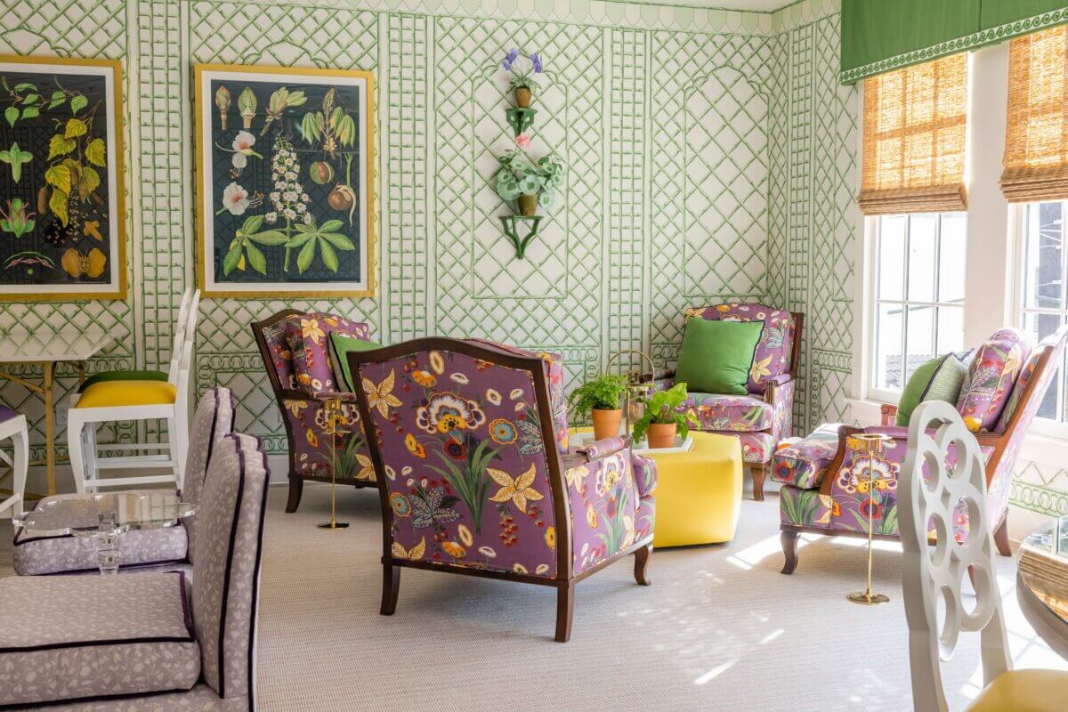 A sunlit sitting room in the Kappa Alpha Theta sorority house features floral-patterned armchairs, green and white lattice wallpaper, potted plants, wall art, and large windows with bamboo shades.