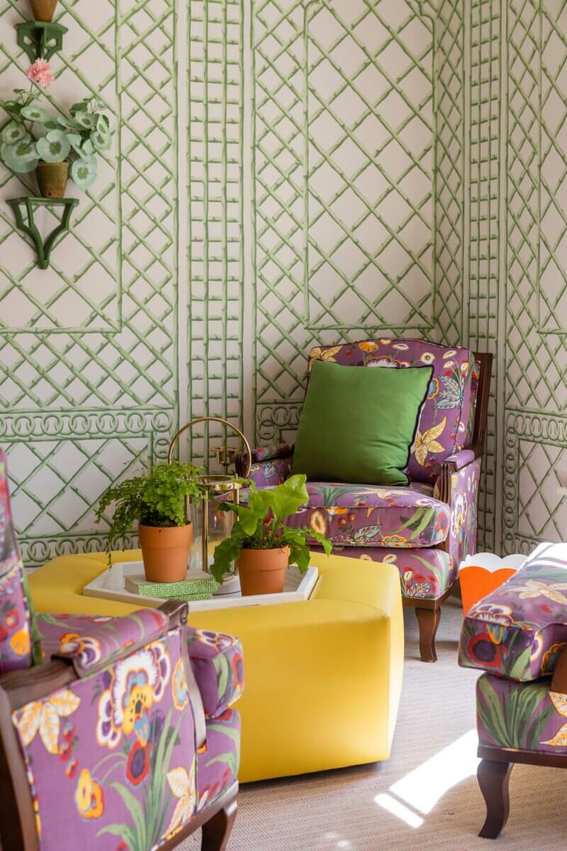 A cozy sitting area in a sorority house, featuring purple floral armchairs, a yellow ottoman, green and yellow pillows, potted plants on a tray, and green lattice-patterned wallpaper—perfect for Kappa Alpha Theta gatherings.