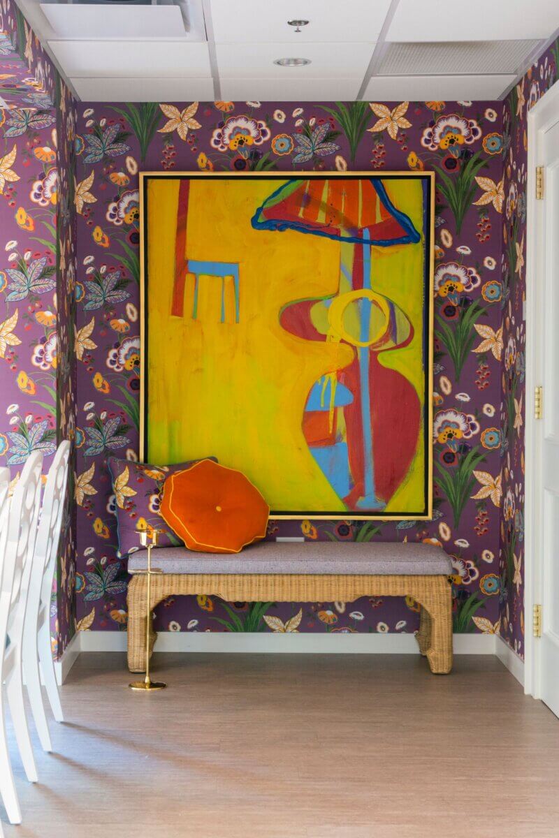 A brightly colored abstract painting hangs above a bench with an orange cushion in a Kappa Alpha Theta sorority house, featuring purple floral wallpaper and light wood flooring.