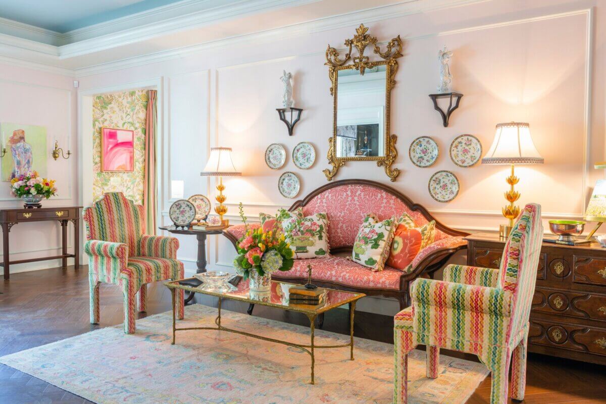 A traditional living room reminiscent of a Kappa Alpha Theta sorority house, with floral-patterned chairs, a wooden sofa, wall-mounted plates, a gold mirror, table lamps, and a glass coffee table with flower arrangements.