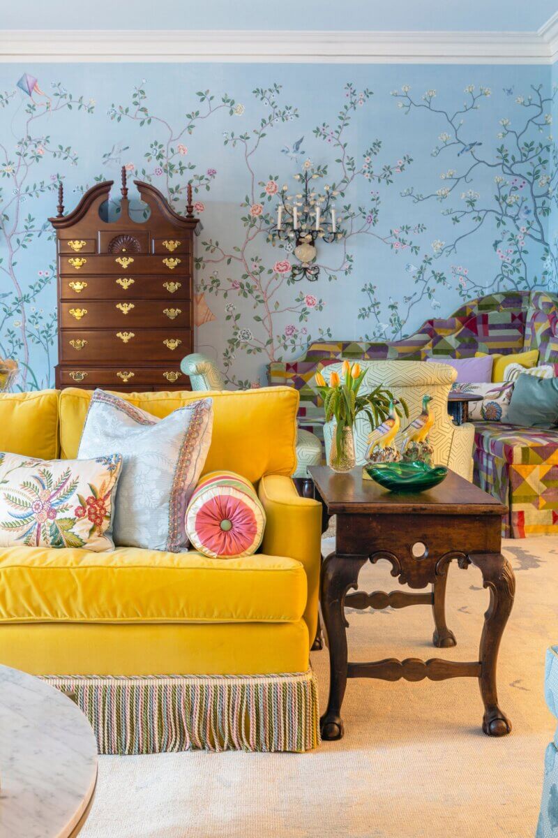 A living room in a Kappa Alpha Theta sorority house with a yellow sofa, floral pillows, a wooden side table, a tall chest of drawers, and a blue wall featuring a painted floral and bird mural.