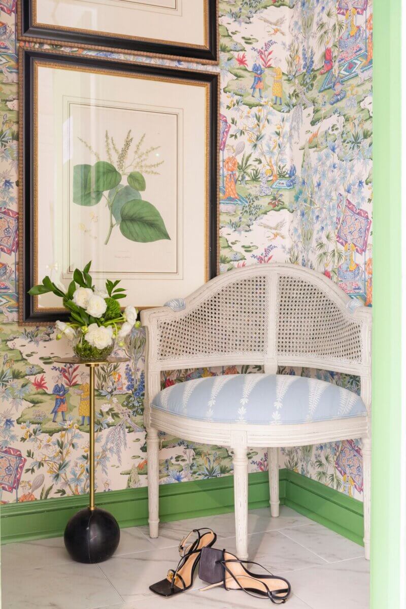 A small white bench with a light blue cushion sits in a corner with floral wallpaper, creating a cozy sorority house vibe, next to a black vase of white flowers and a pair of high-heeled shoes on the floor.