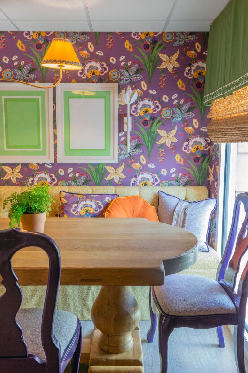 A cozy dining nook in a Kappa Alpha Theta sorority house with a wooden table, cushioned chairs, bright floral wallpaper, framed wall art, and a potted plant on the table.